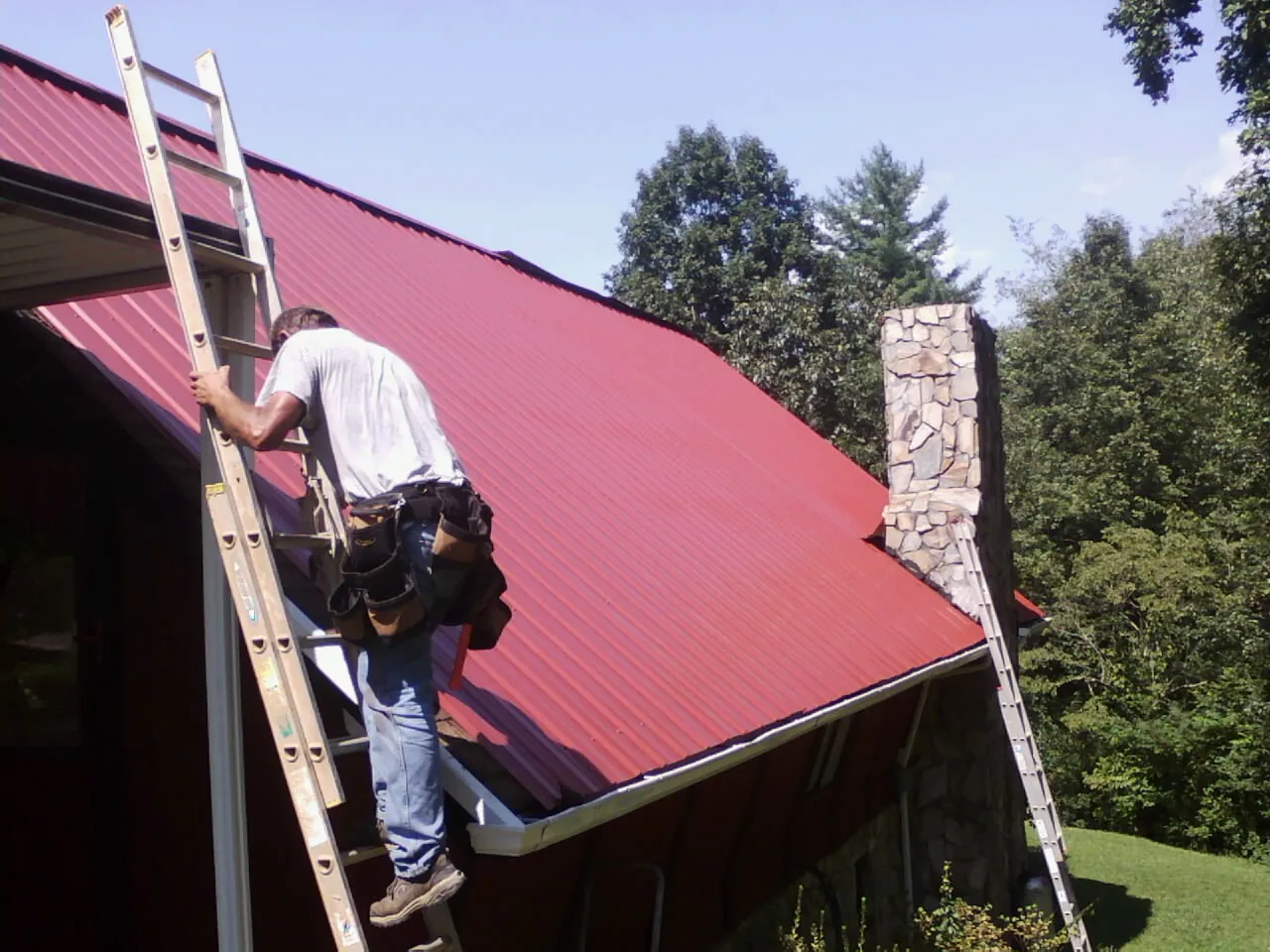 Roofer on a ladder beside a red metal roof and stone chimney — Soffit & Fascia Repair by Roofing Brothers in Geneva